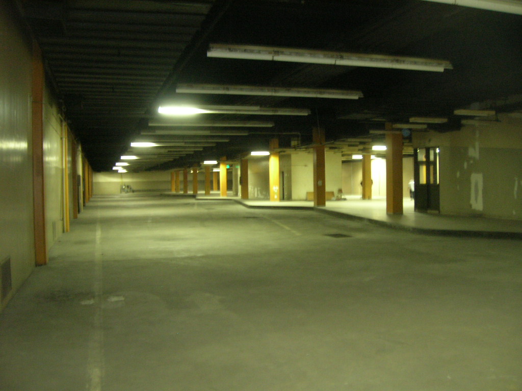 color photo showing a concrete curb forming a sawtooth shape, in a basement structure supported by yellow concrete columns and lit by fluorescents. Elevators and stairways extend down from the next level of the building.