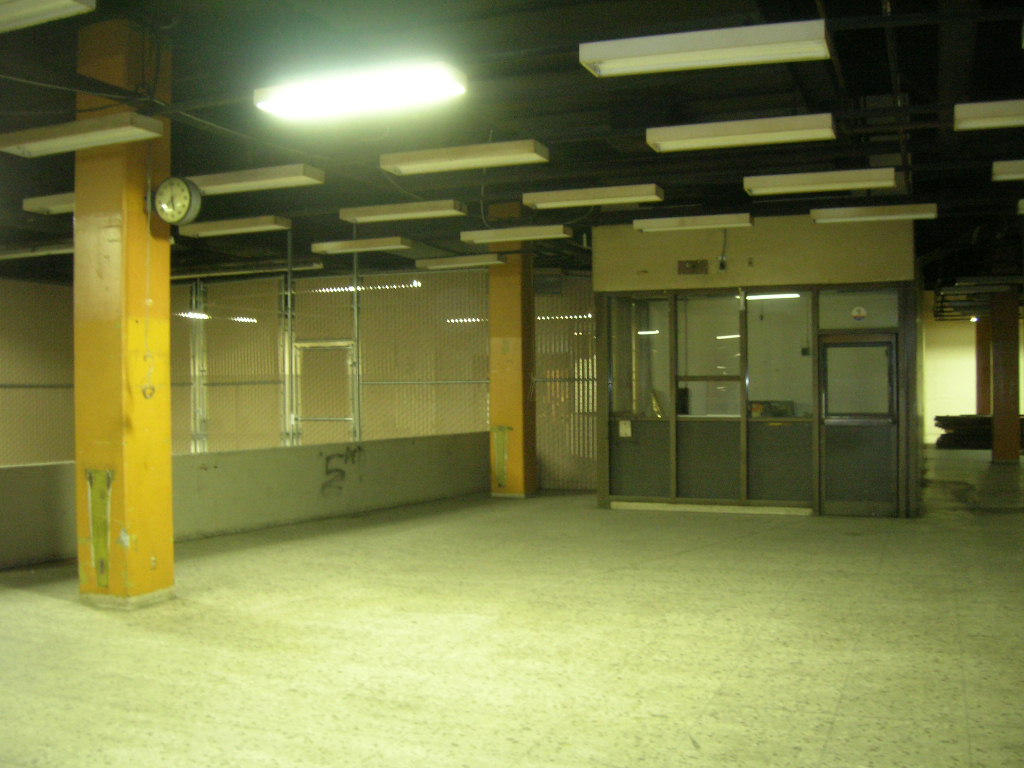 color photo showing a metal framed glass booth, somewhere along the concrete boarding platform. There is a plastic-slatted chain link fence next to it, and a clock still affixed to one of the yellow columns.