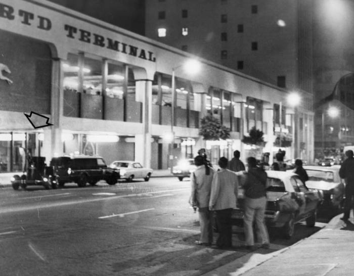 Black and white photo of a crowd od people standing across the street from the entrance of the bus terminal. a car with a bulbous trailer is noted by an arrow, just outdise the entrance.