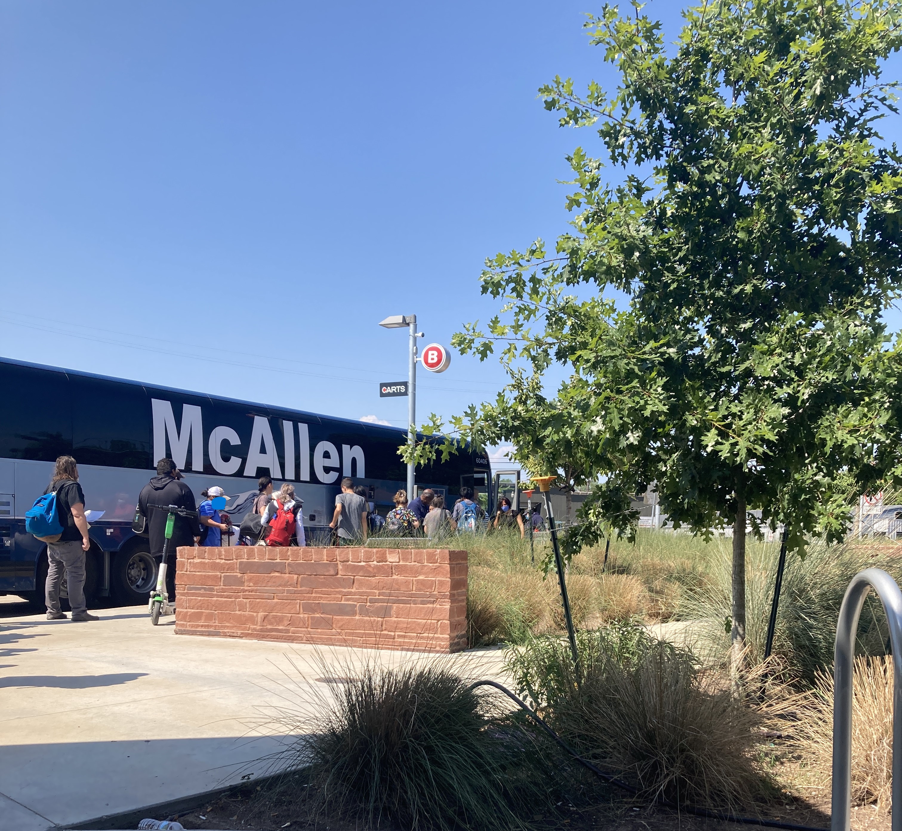 passenger line up to board a large gray and blue coach bus with the name McAllen emblazoned on its windows 