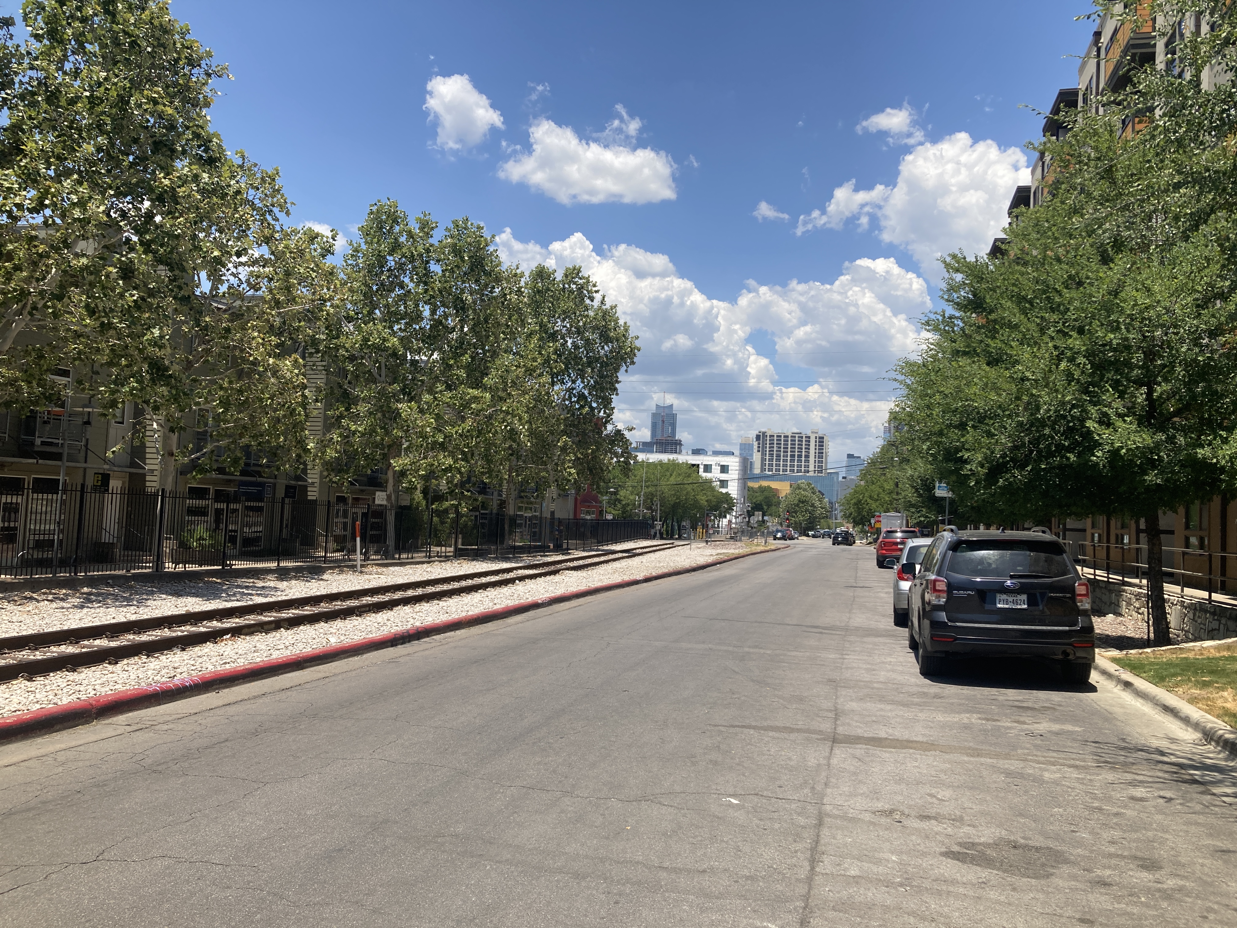 a gravel-ballasted train track separated from a street by a curb. on either side are shade trees and mid rise buildings, with taller buildings in the distance 