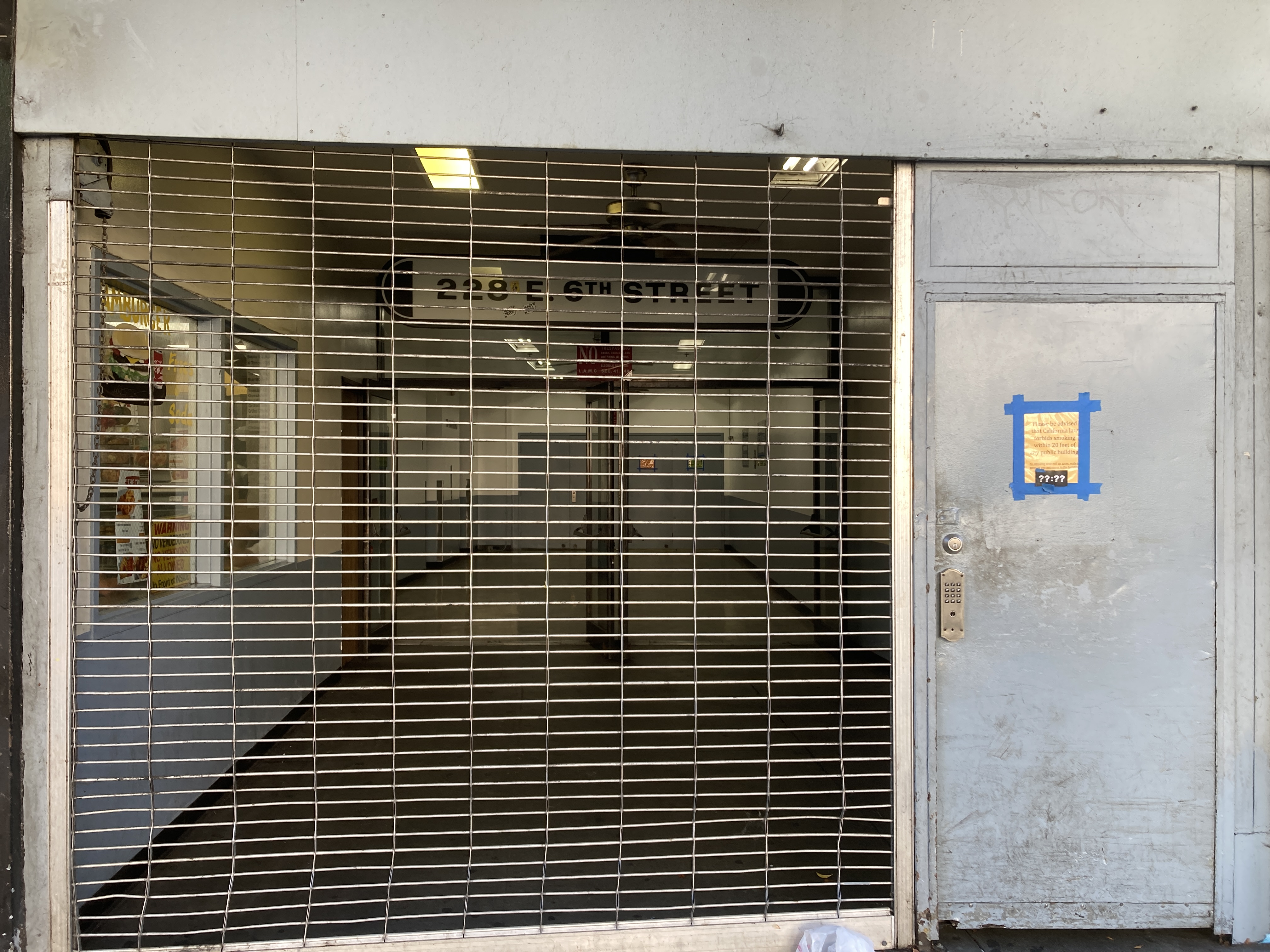 an alcove in the facade of the building is enclosed by a roll-down grated gate, through which can be seen four blue metal doors and a lozenge-shaped sign reading 228 E 6TH STREET