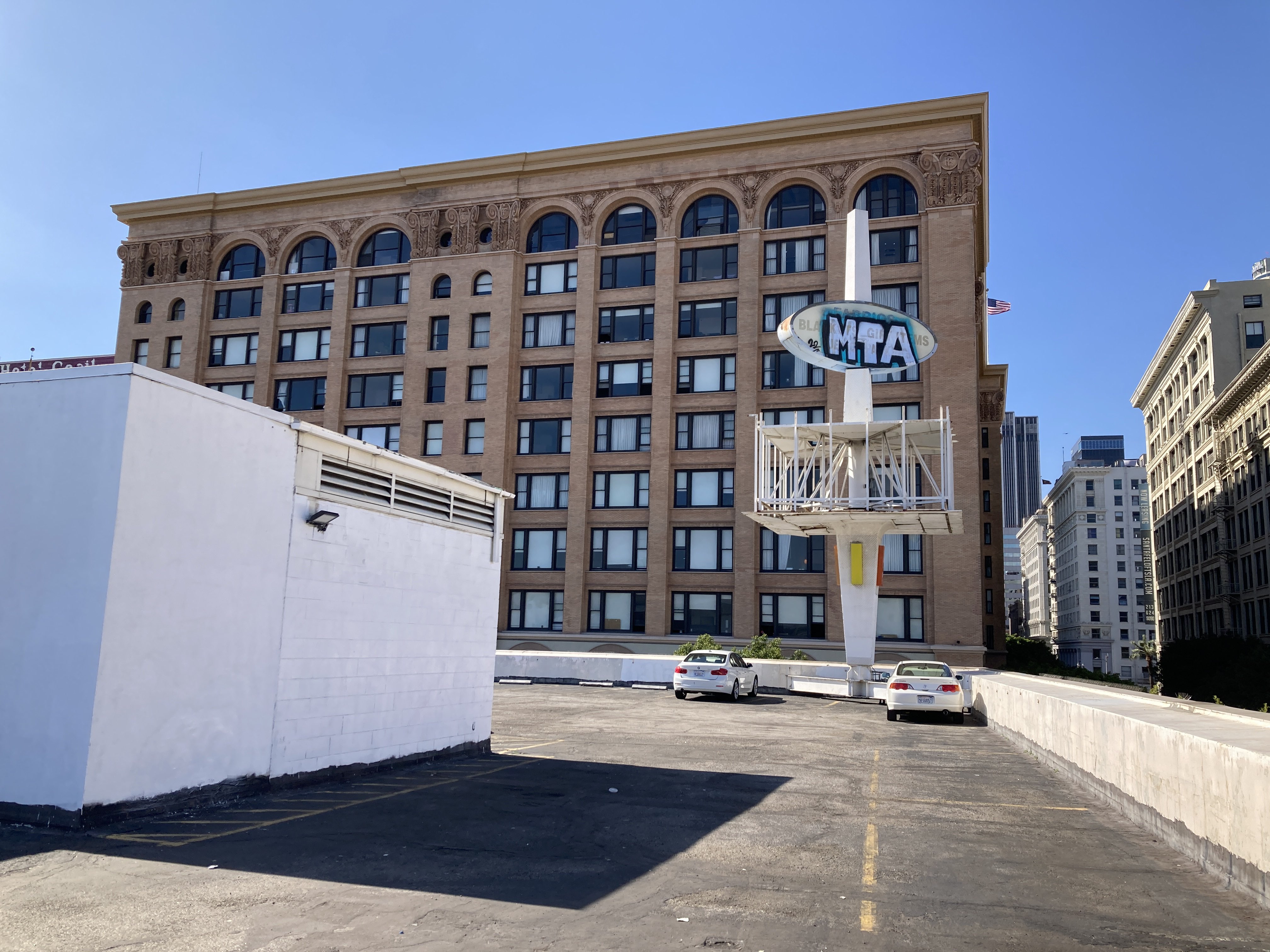 the large multifaceted sign on the rooftop of the building at sixth and los angeles. it has a rotating oval on top and a rectangle on each side. cars are parked nearby