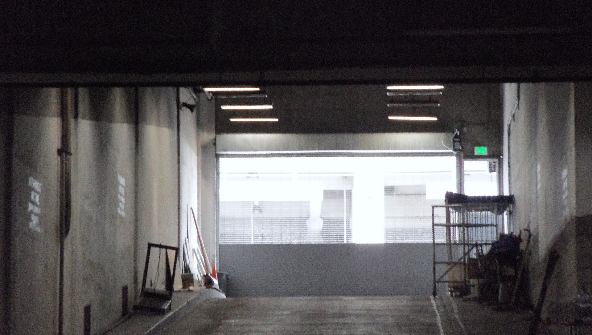 color photo showing a concrete ramp up to street level, surrounded by shelves and pieces of fence. A parking structure is visible across the street outside its roll-down gate.