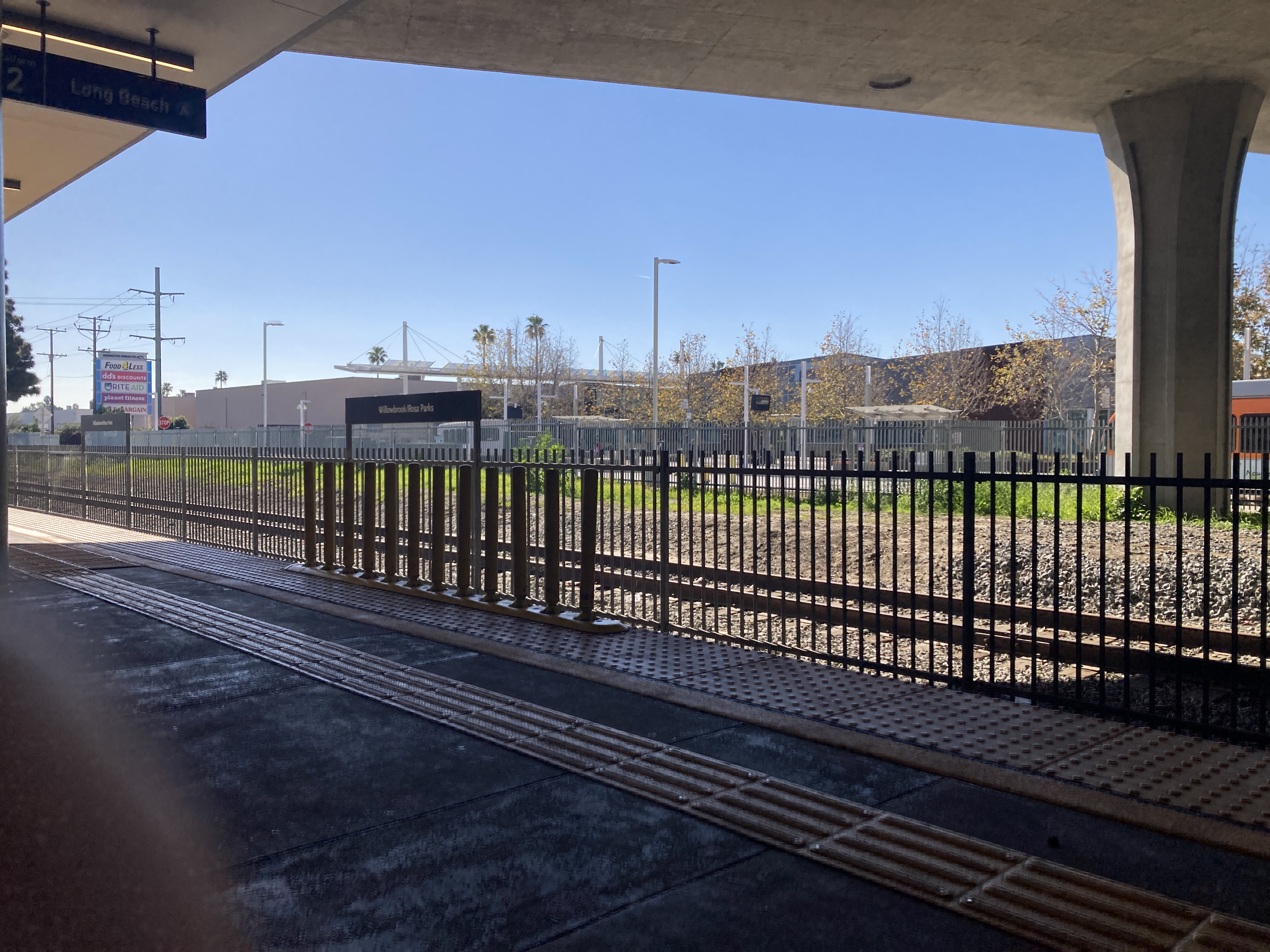 across two standard gauge train tracks separated by a fence, buses stop in front of shade and light canopies.