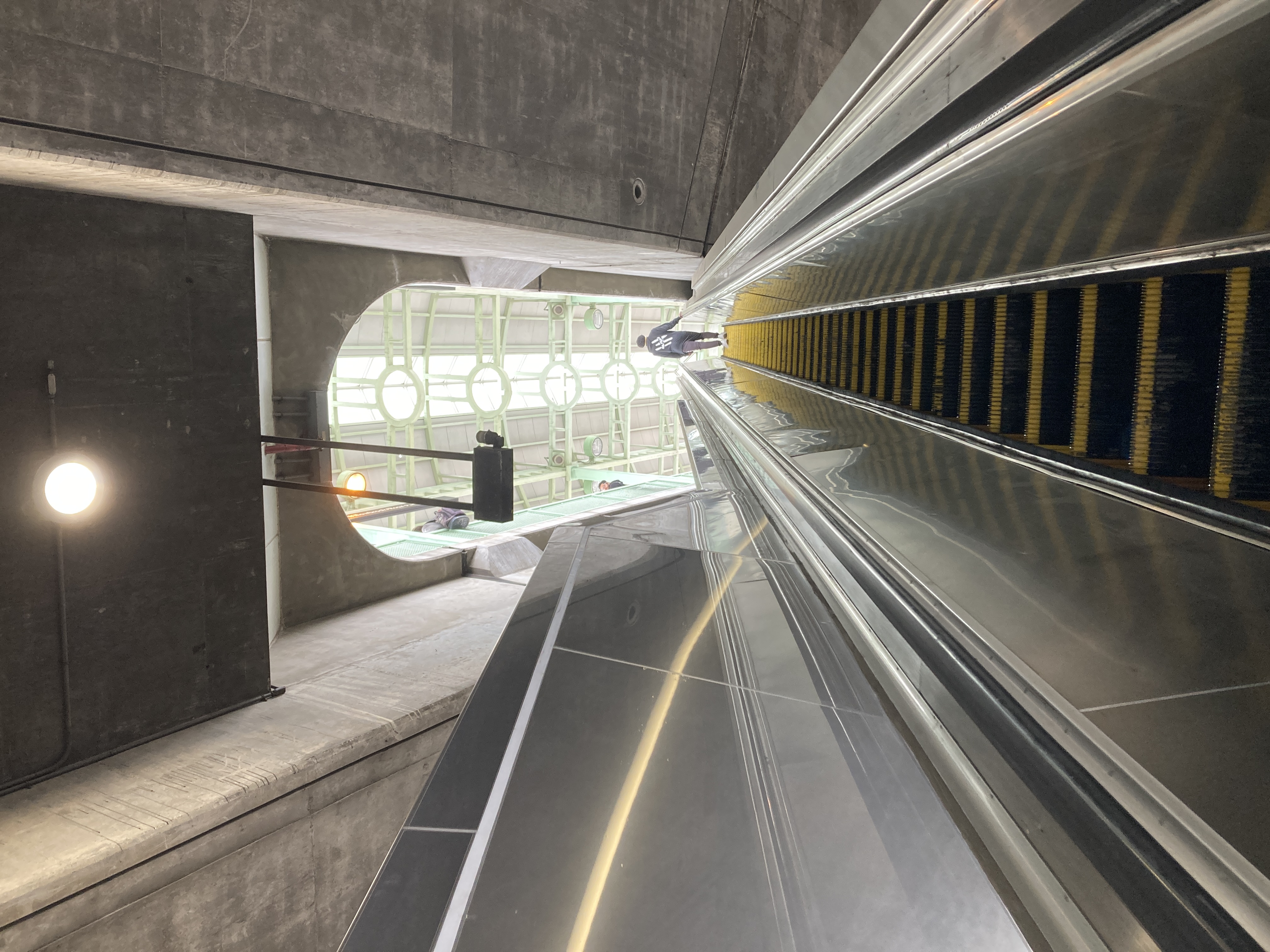 looking up an escalator through a lozenge shaped opening in the viaduct. the metal canopy and plexiglass skylight of the green line platform let sunlight in from above.